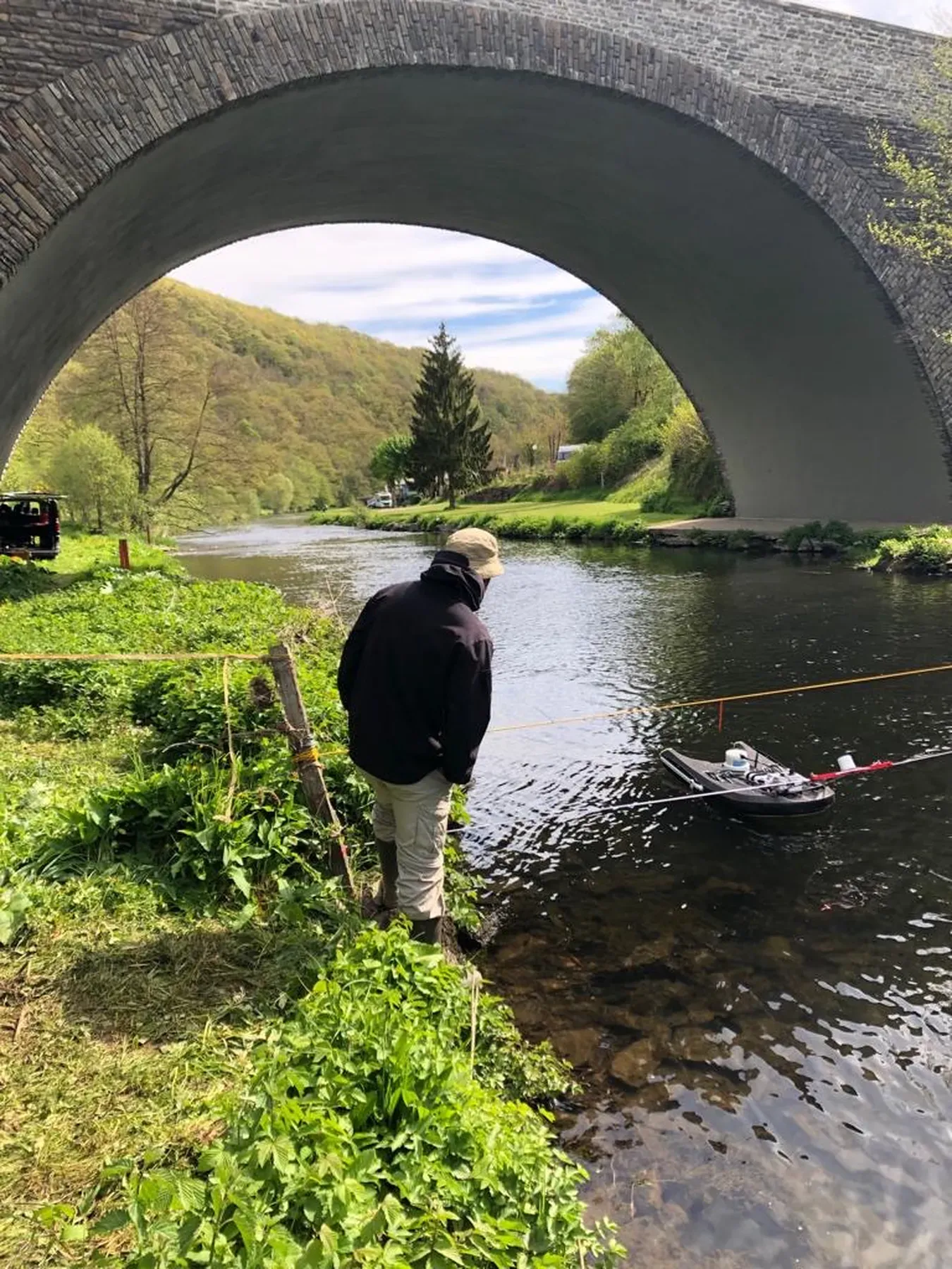 Remote-control bathymetric survey boat measuring river bed levels beneath a stone arch bridge.