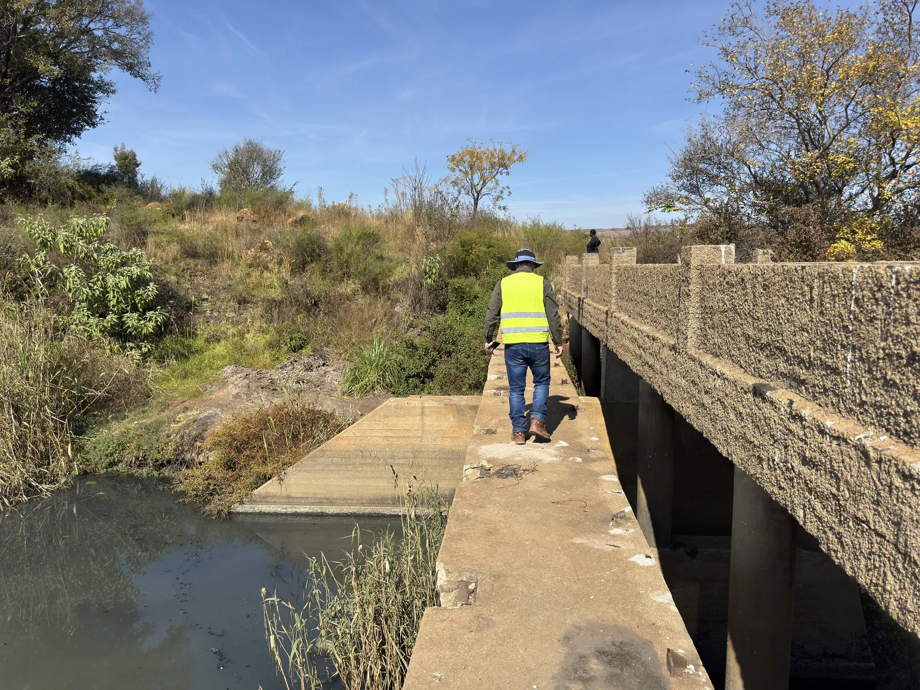 Engineer in hi-vis walking a weir wall beside a concrete road bridge during a hydraulic inspection.