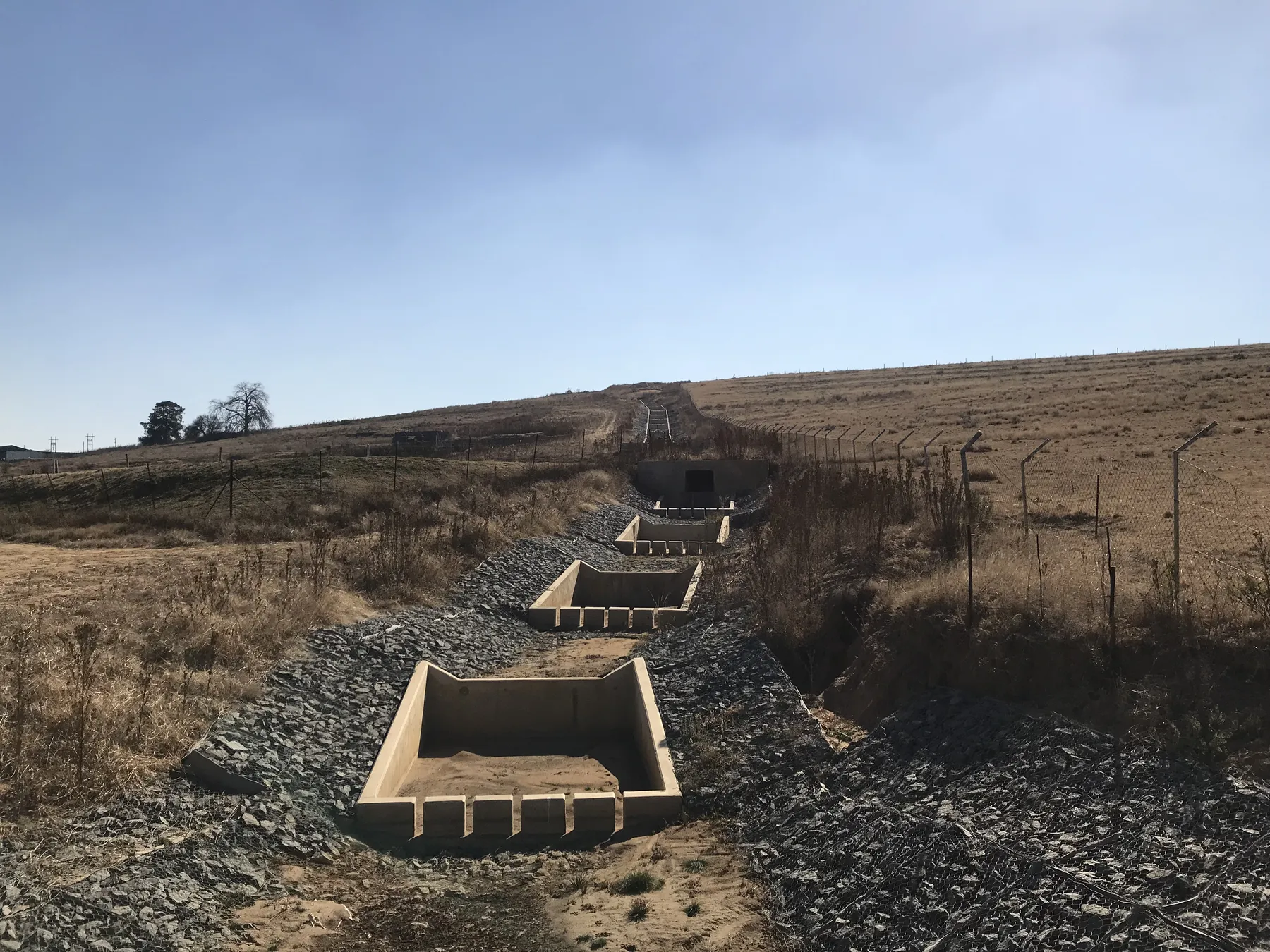 Stepped concrete energy dissipator structures descending a steep grassed slope in open veld.