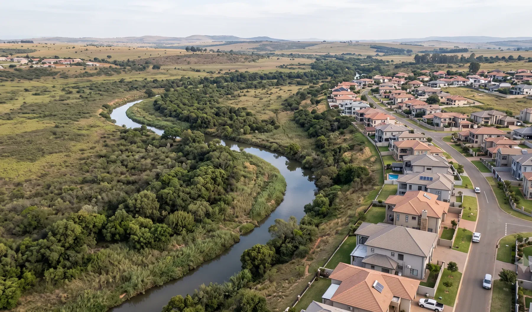 Aerial view of a river floodplain bordering a residential development, showing the natural watercourse and adjacent low-lying land.