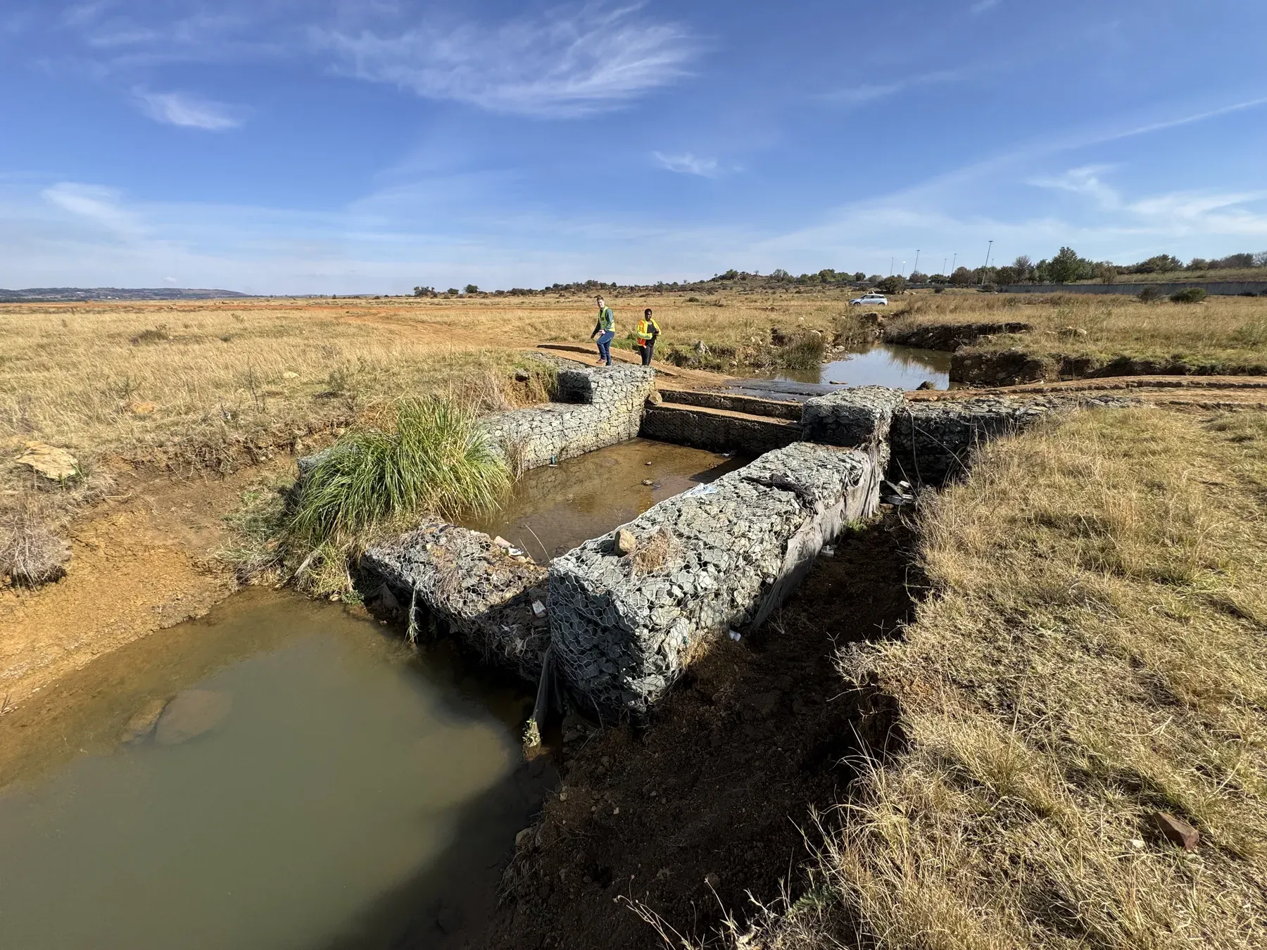 A failed gabion structure with burst baskets and displaced rock along a watercourse.